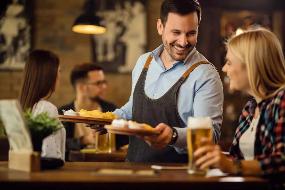 A friendly waiter is bringing food to a table in a pub and smiles at one of the guests