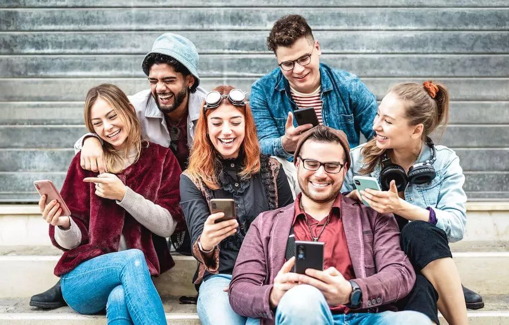 Mozrest - Group of friends seating in the street smiling and laughing while looking at social media on their smartphone