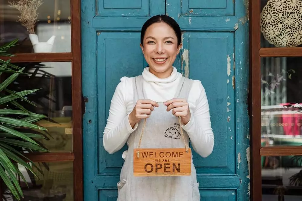 Mozrest - A smiling restaurant manager is standing in front of her blue restaurant door with a welcome sign that reads 'we are open'.
