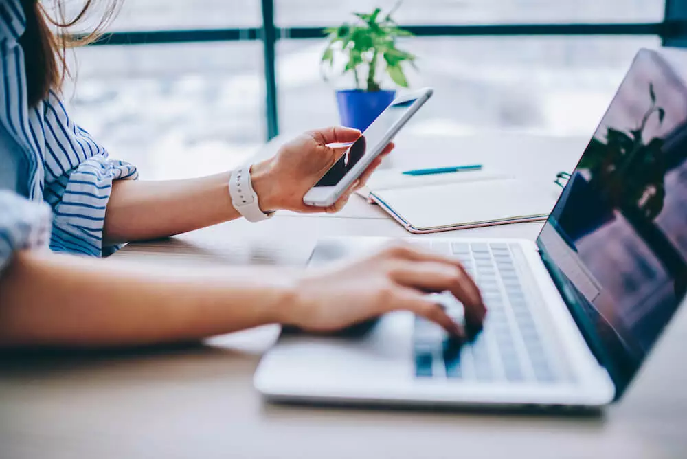 Mozrest - Woman using her smartphone while working on a laptop at her desk in the office