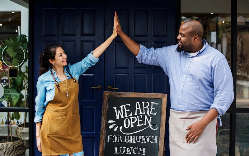 Two restaurant managers, a woman and a man, smiling at each other and doing a high five in front of the main door as they open their restaurant for brunch, lunch and dinner.