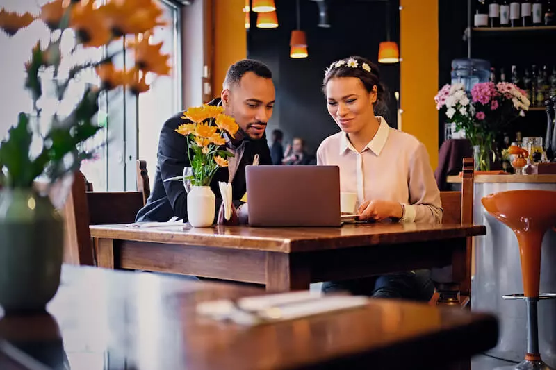 Un homme et une femme assis à une table dans un restaurant clair et lumineux, gèrent facilement la réputation en ligne de leur établissement sur un ordinateur portable grâce à Listing & Review.