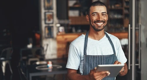 Le responsable du restaurant se tient devant son restaurant, souriant et utilisant une tablette pour vérifier ses réservations dans son logiciel de réservation.