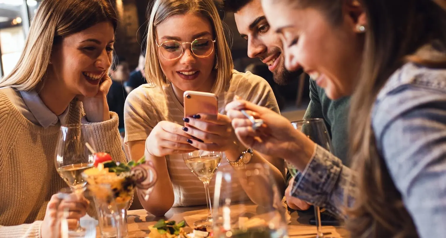 Mozrest - Solutions for Booking Channels - Group of friends seating at a table in a restaurant searching for a their next place to eat on their mobile phone