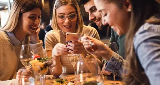  - Group of friends seating at a table in a restaurant searching for a their next place to eat on their mobile phone